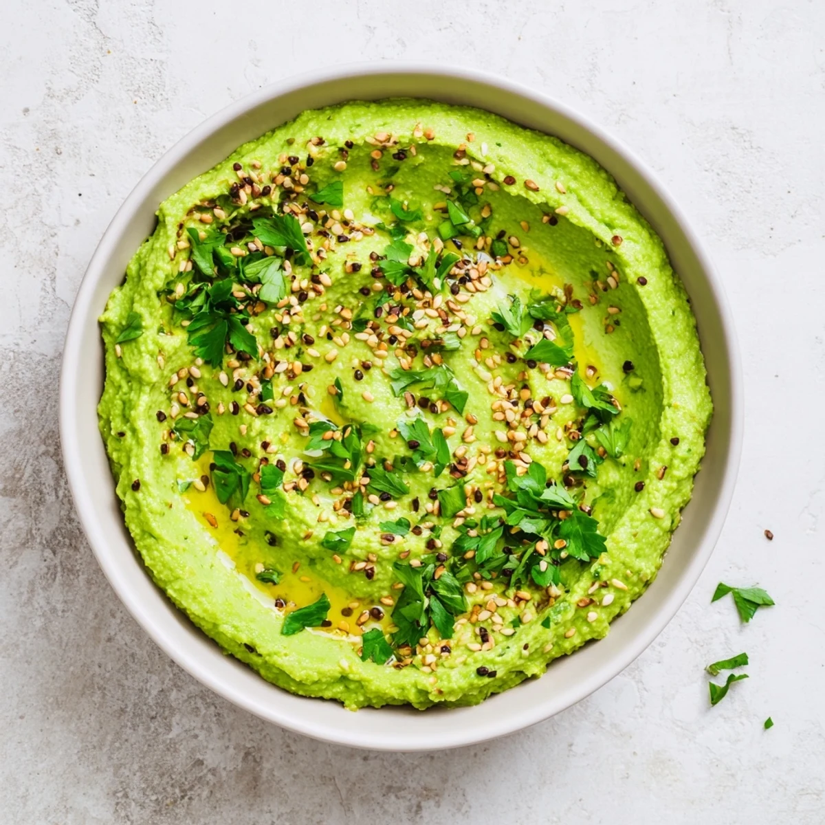 Creamy green edamame hummus in a bowl, garnished with olive oil, toasted sesame seeds, and fresh parsley leaves.  