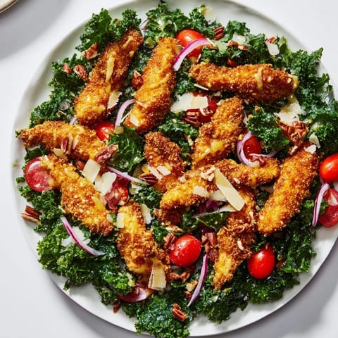 Golden, crispy chicken strips rest on a vibrant bed of kale salad with tomatoes and onions, drizzled with tangy maple-Dijon dressing.  
